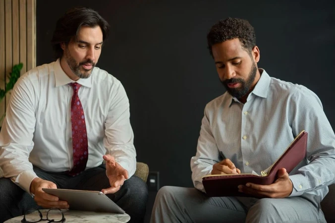 Two business professionals reviewing a tablet and taking notes in a modern office setting, dressed in button-down shirts and ties, indoors.